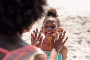 A joyful girl gets sunscreen applied by her mom on a sunny beach day.